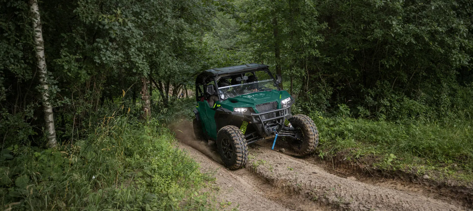 Front end view of a Polaris GENERAL XP 1000 side-by-side with headlights illuminated driving on a forest trail.