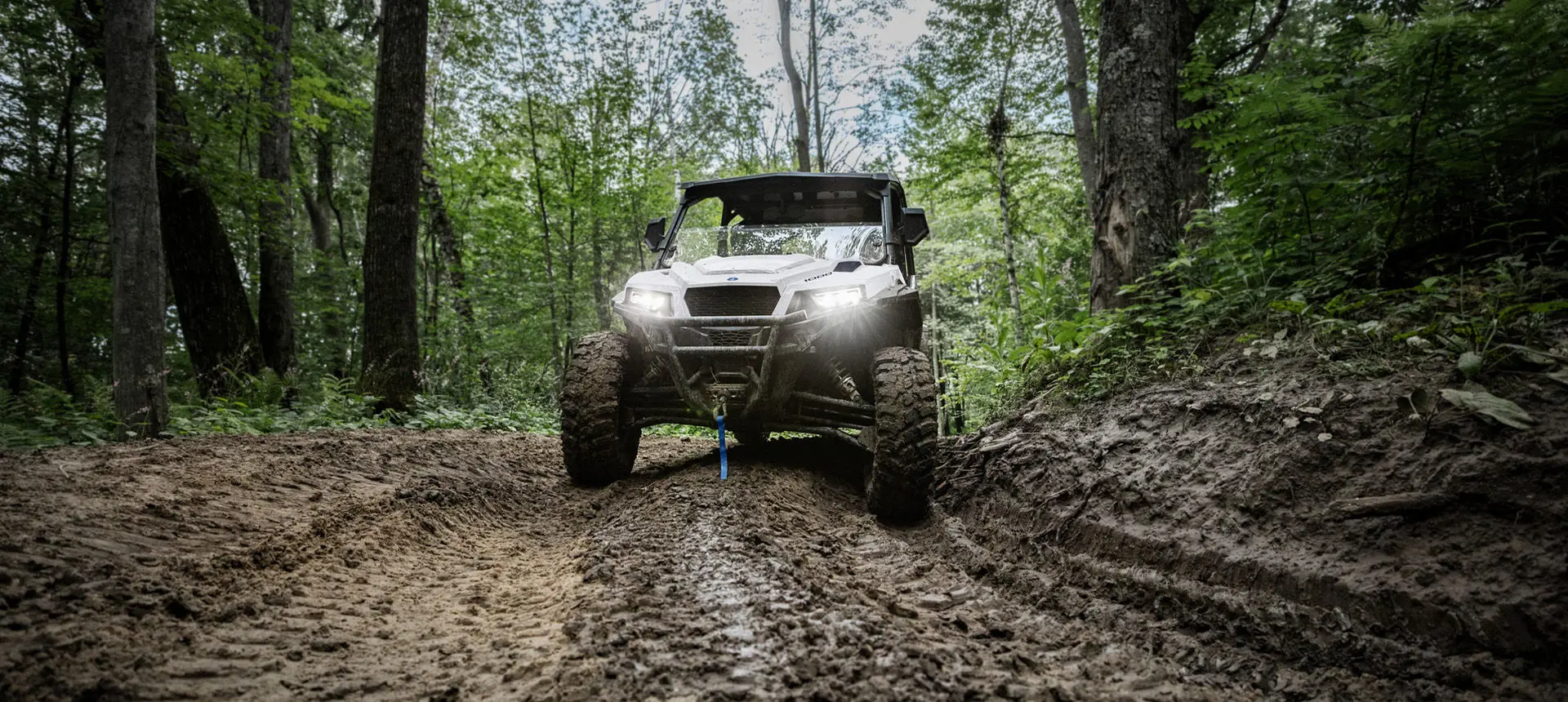 Front end view of a Polaris GENERAL XP 1000 side-by-side driving on a muddy trail in the forest.