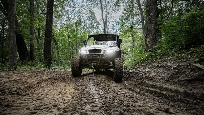 Front end view of a Polaris GENERAL XP 1000 side-by-side driving on a muddy trail in the forest.