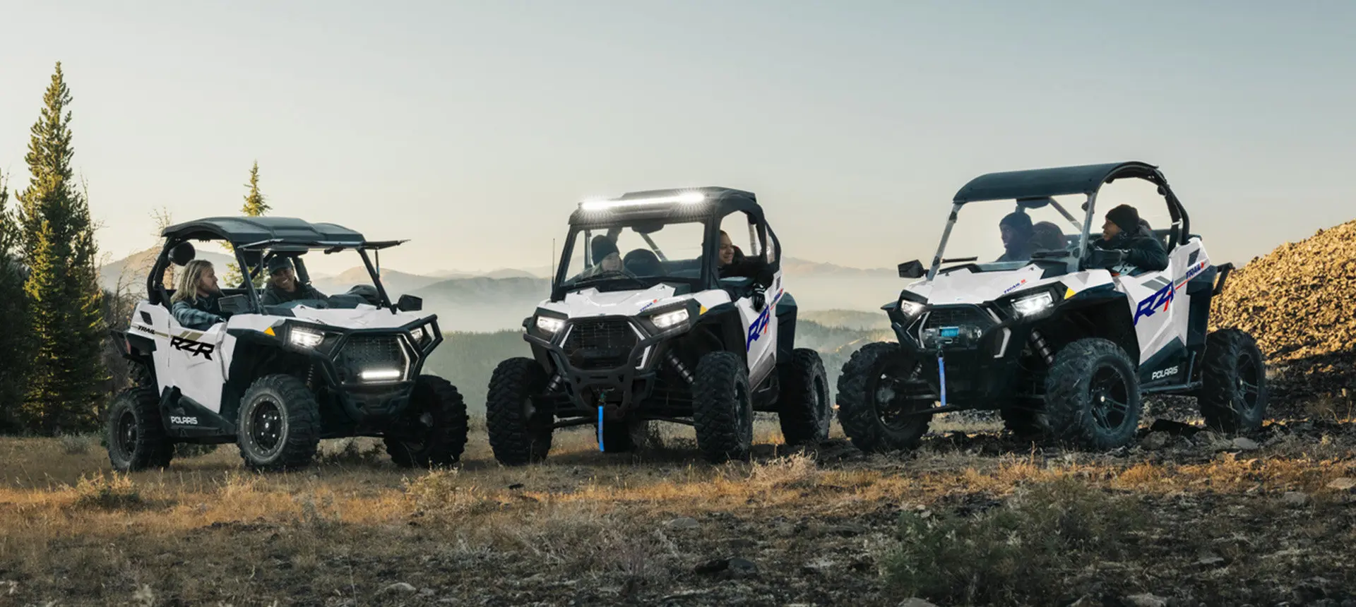 Three Polaris RZR Trail side-by-sides parked in a lineup with illuminated lights.