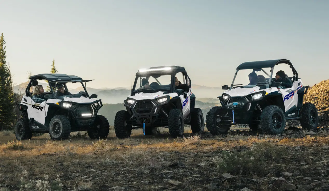 Three Polaris RZR Trail side-by-sides parked in a lineup with illuminated lights.