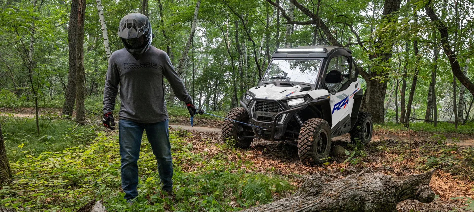 A person pulling the winch out of the front end of a Polaris RZR Trail side-by-side in the forest.