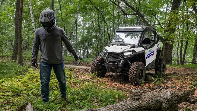 A person pulling the winch out of the front end of a Polaris RZR Trail side-by-side in the forest.