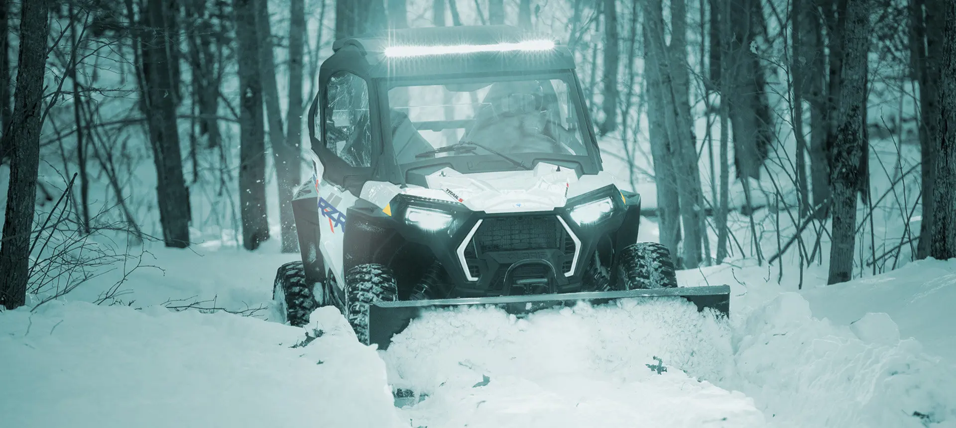 A person in a Polaris RZR Trail side-by-side plowing snow in the winter.