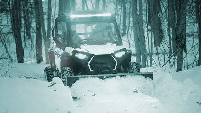 A person in a Polaris RZR Trail side-by-side plowing snow in the winter.