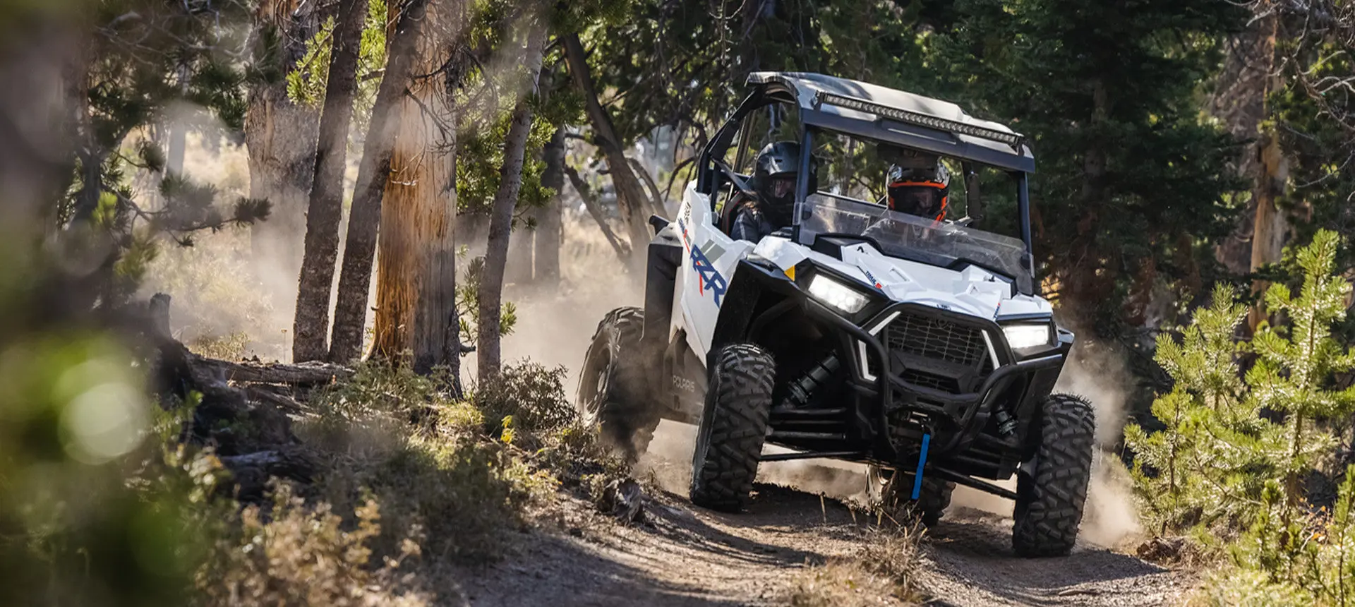 Front end view of a Polaris RZR Trail side-by-side with lights on on a trail.