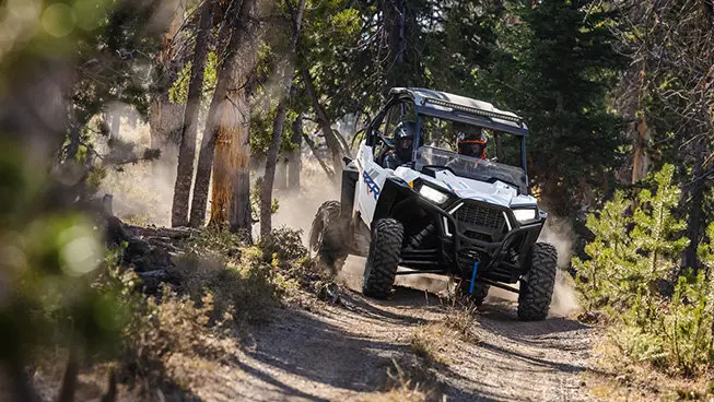 Front end view of a Polaris RZR Trail side-by-side with lights on on a trail.