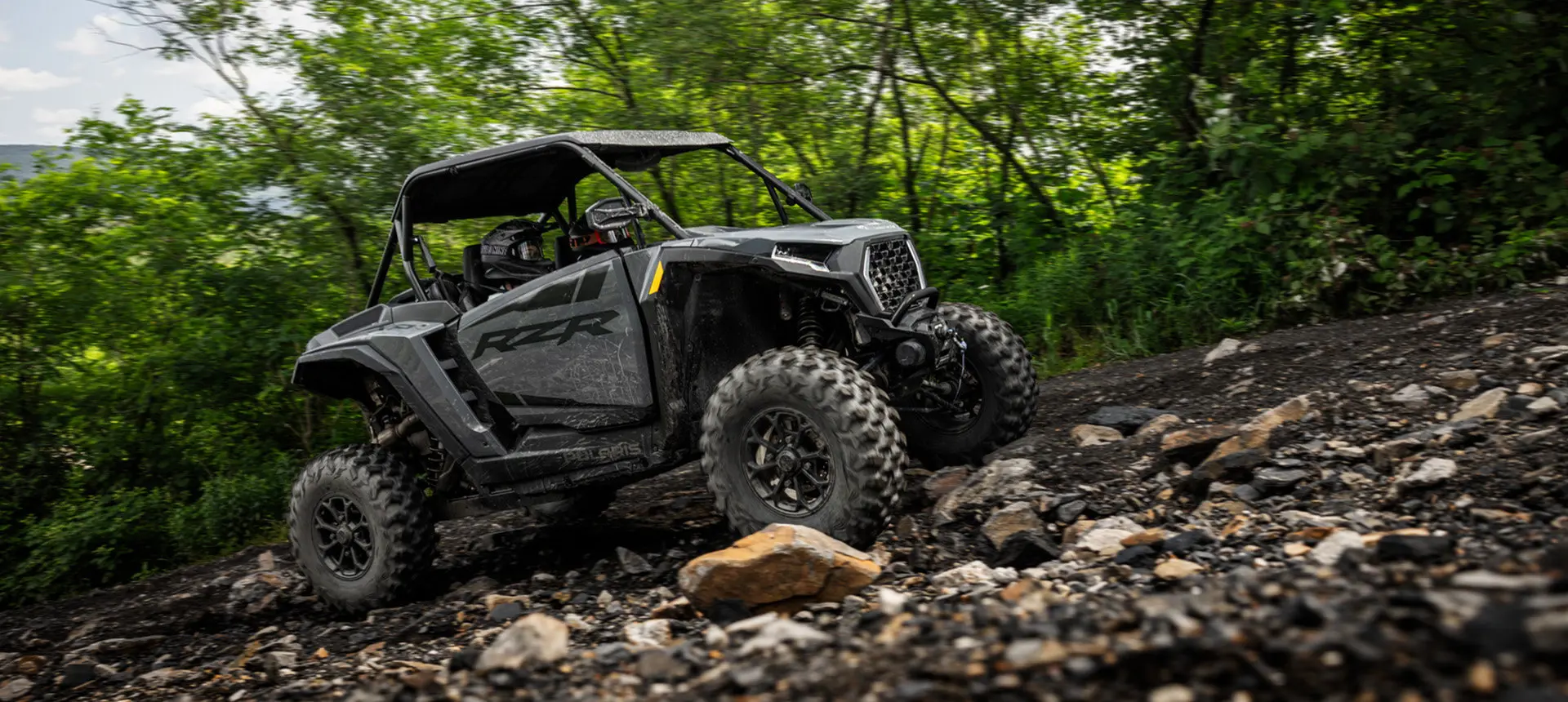 Side profile of 2 riders in a Polaris RZR XP traveling over rocky terrain on a forest trail.