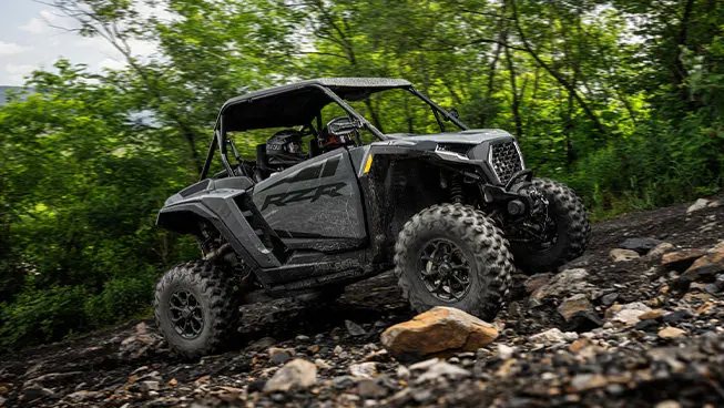 Side profile of 2 riders in a Polaris RZR XP traveling over rocky terrain on a forest trail.