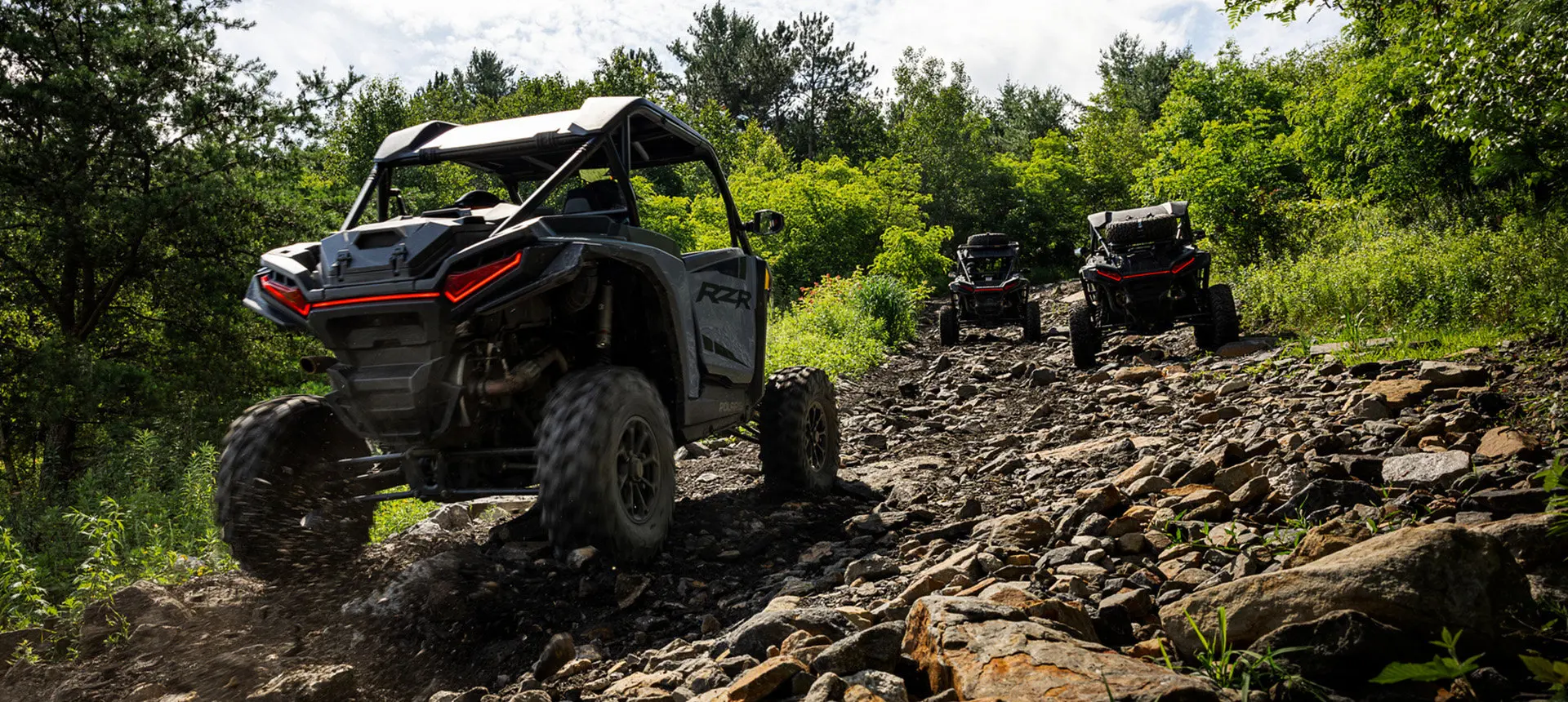 Three Polaris RZR XP side-by-sides driving on a rocky trail.