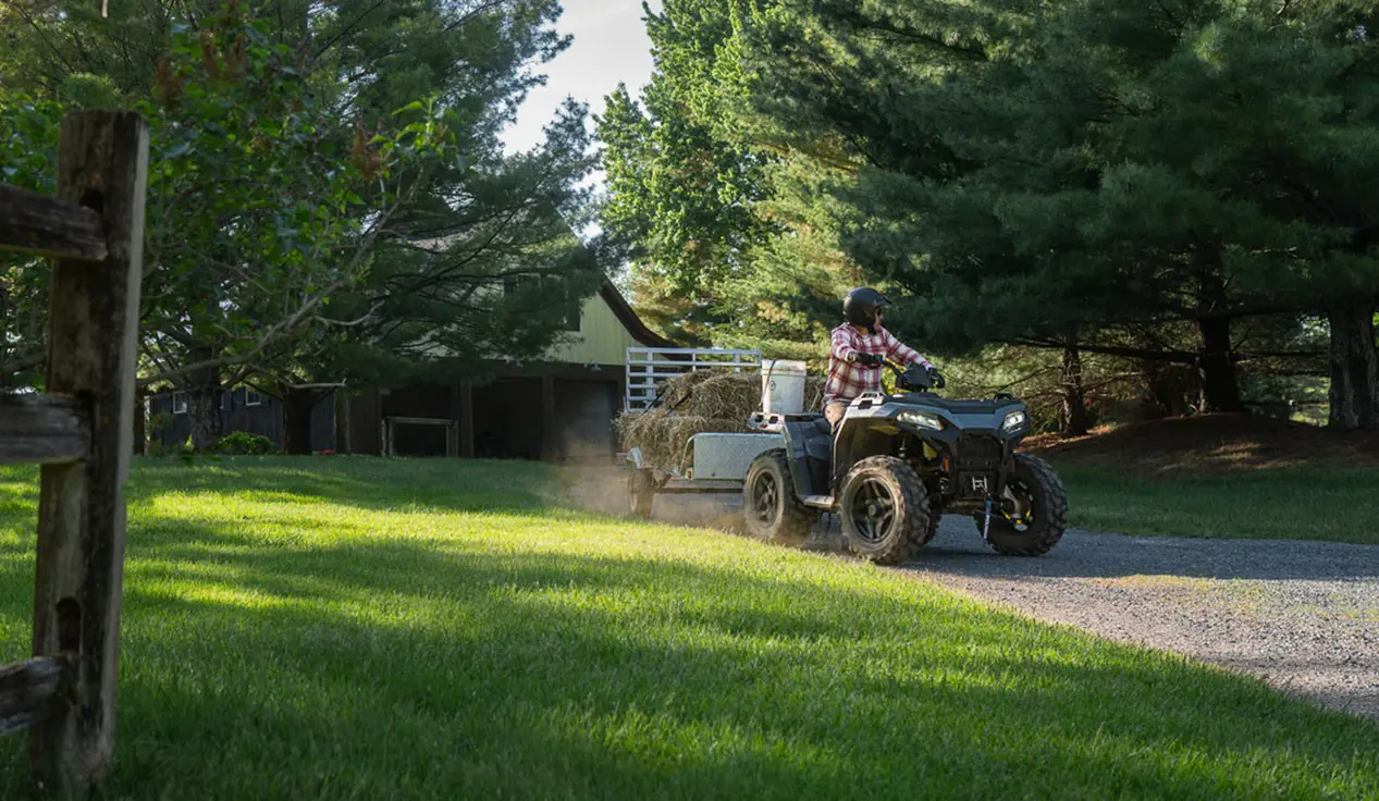 A rider on a Polaris Sportsman 500 ATV pulling a trailer loaded with hay bales.