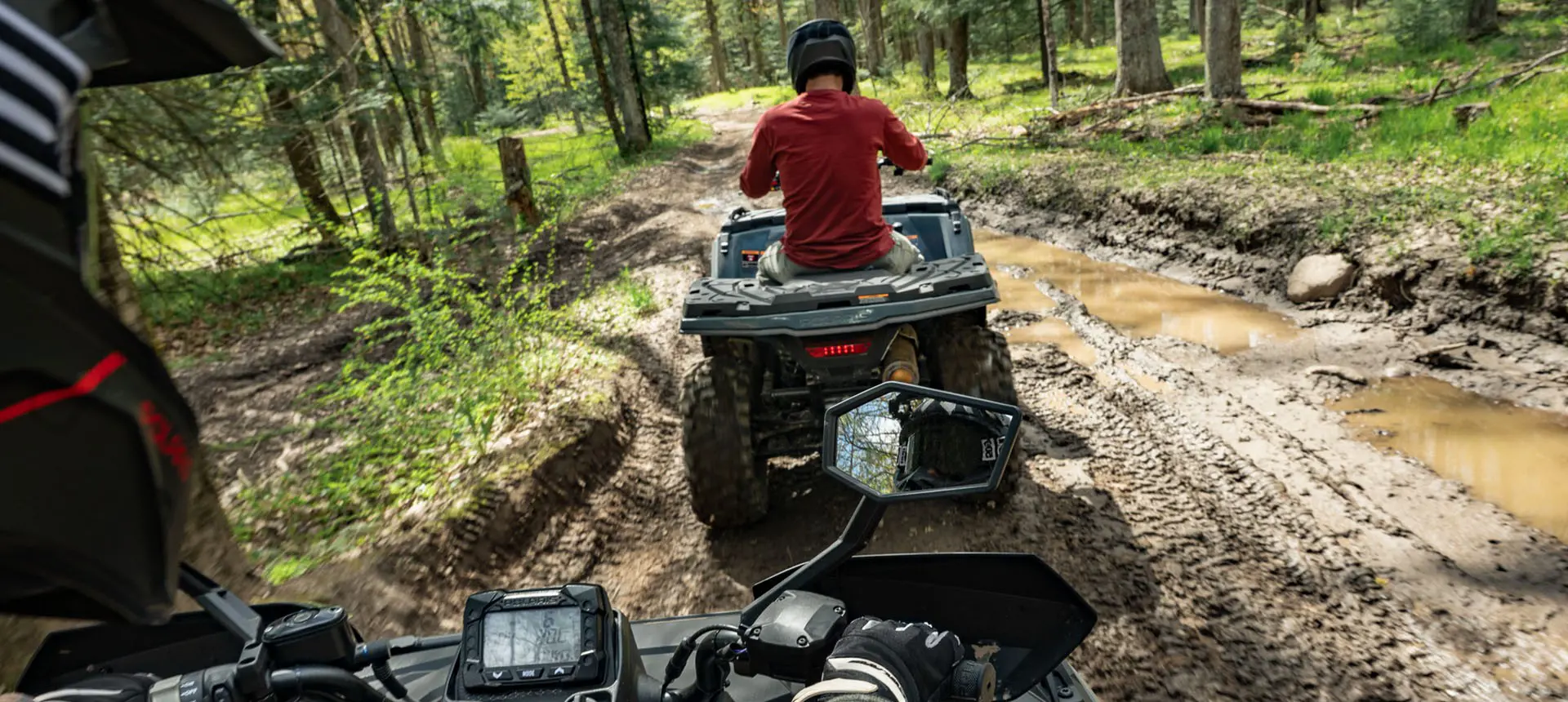 A rider on a Polaris Sportsman 500 ATV with side view mirrors driving behind another Polaris Sportsman 500 ATV on a muddy trail.