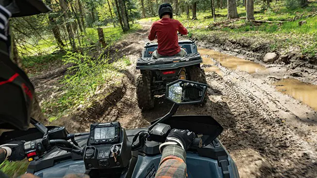 A rider on a Polaris Sportsman 500 ATV with side view mirrors driving behind another Polaris Sportsman 500 ATV on a muddy trail.