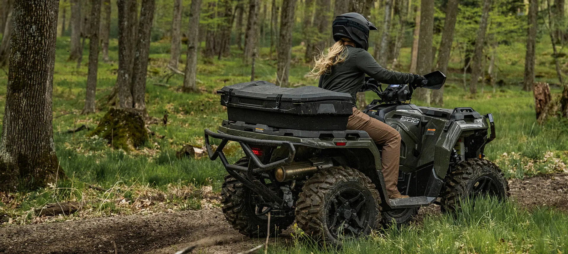 A rider on a Polaris Sportsman 500 ATV with a rear cargo box accessory driving on a trail in the forest.