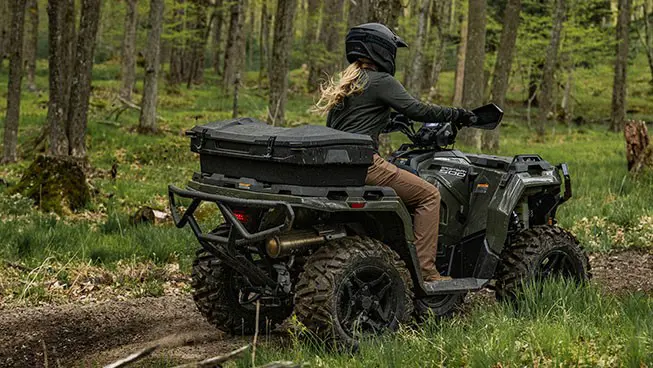 A rider on a Polaris Sportsman 500 ATV with a rear cargo box accessory driving on a trail in the forest.
