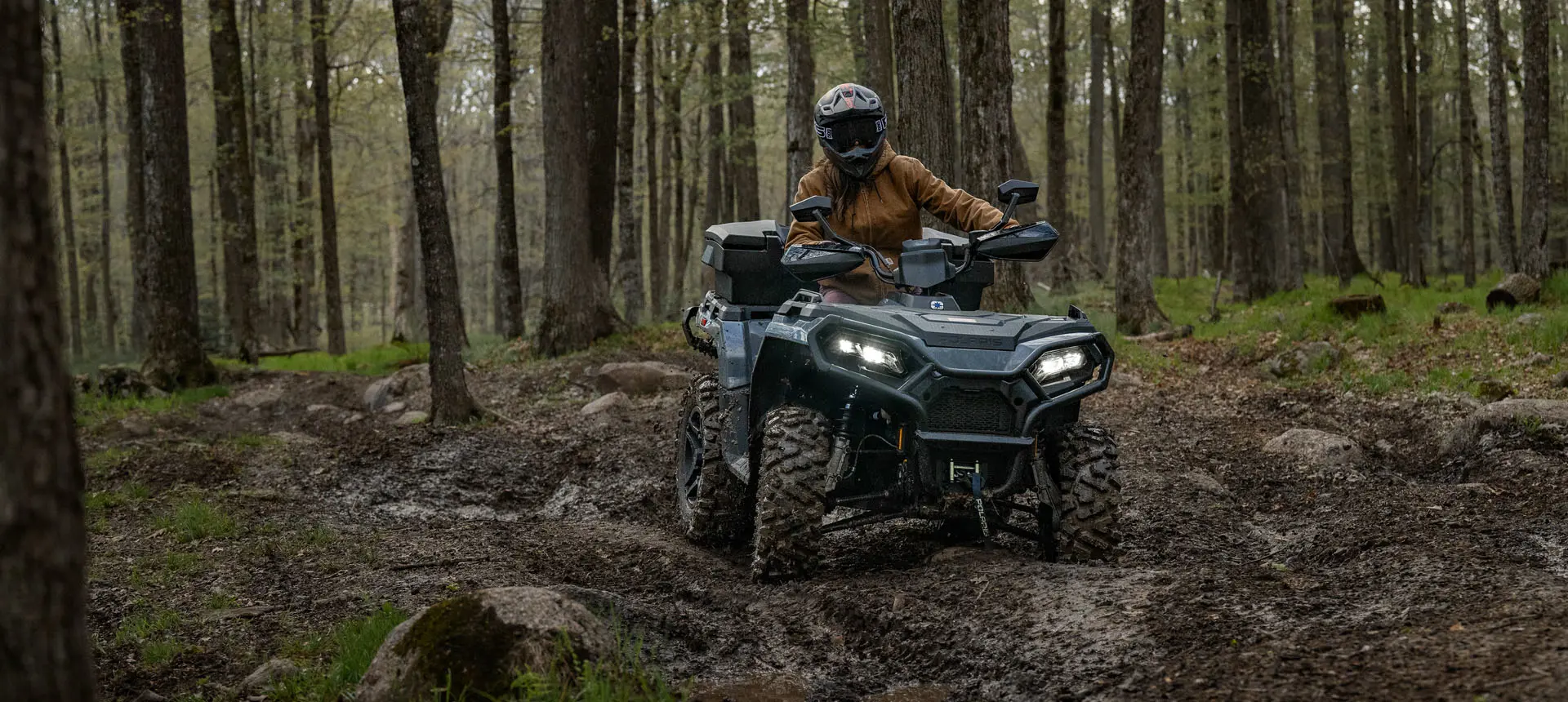 A Polaris Sportsman 500 ATV driving in the forest over a rocky uneven trail.