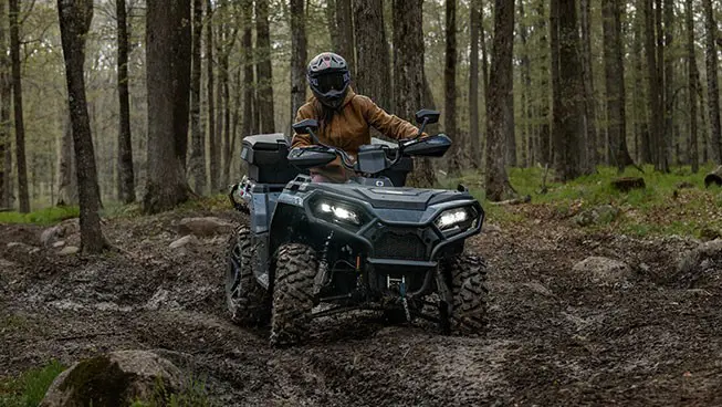 A Polaris Sportsman 500 ATV driving in the forest over a rocky uneven trail.