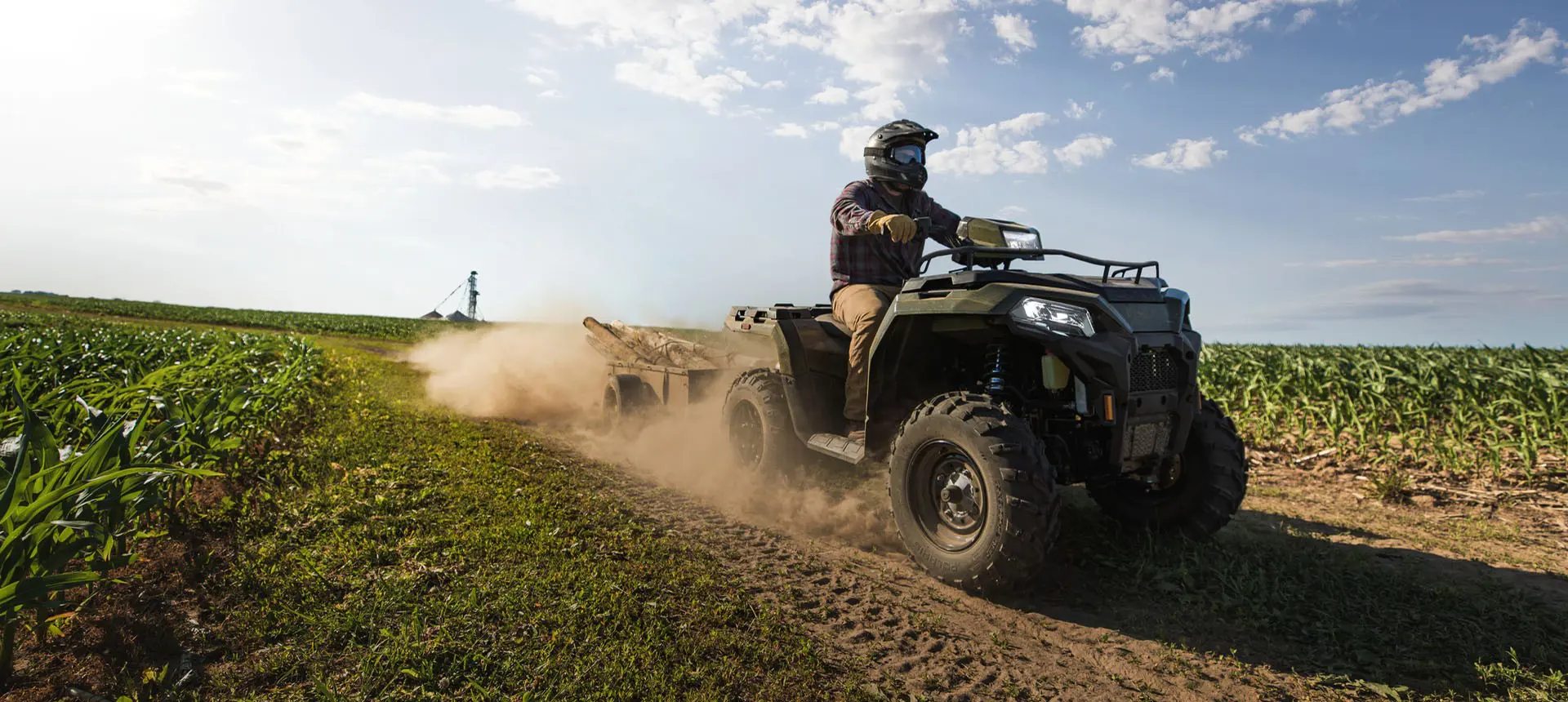 A rider on a Polaris Sportsman 600 ATV pulling a trailer on a country road near crops.