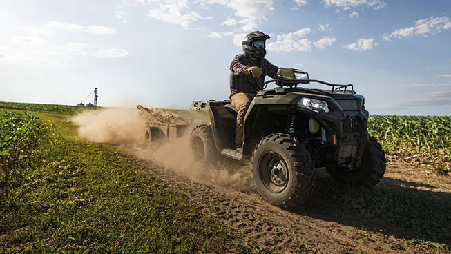 A rider on a Polaris Sportsman 600 ATV pulling a trailer on a country road near crops.