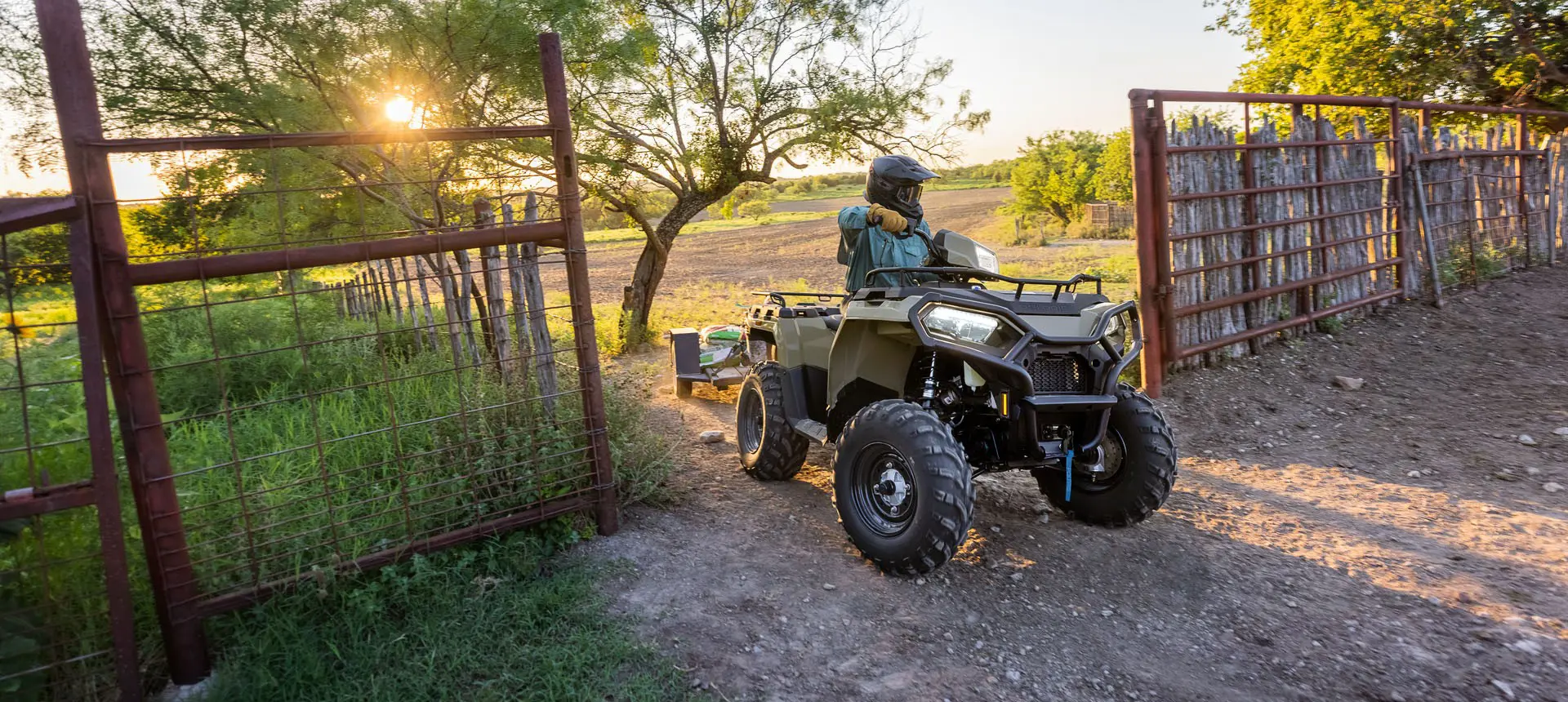 Rider on a Polaris Sportsman 600 ATV pulling a trailer through a gate.