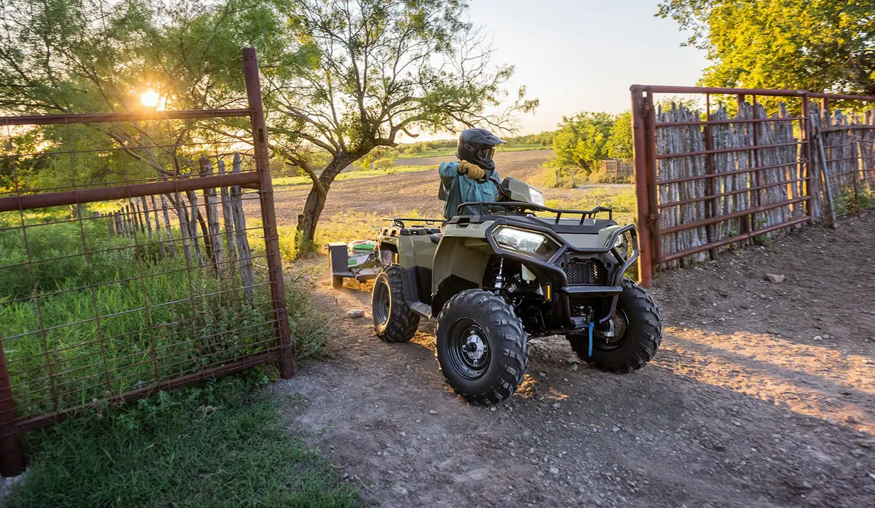 Rider on a Polaris Sportsman 600 ATV pulling a trailer through a gate.