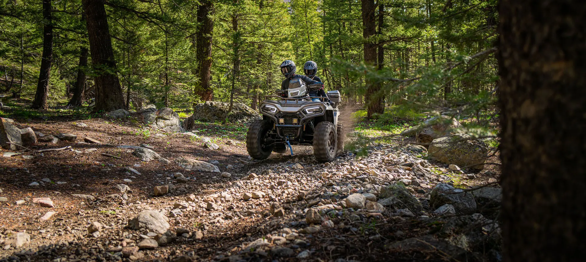 2 riders on a Polaris Sportsman 600 Touring ATV driving through the forest.