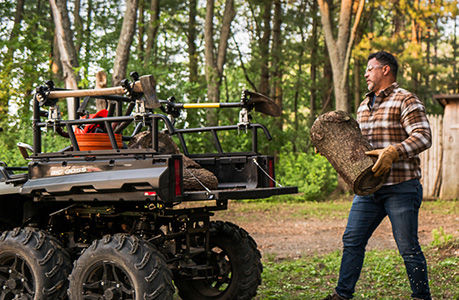 A man loading firewood into the cargo box of a Polaris Sportsman 6x6 600 ATV with racks and grip accessories installed.