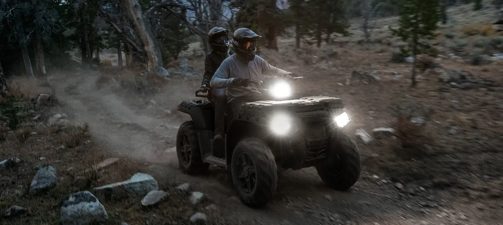 Two riders on a Polaris Sportsman Touring 850 ATV with illuminated headlights on a trail.
