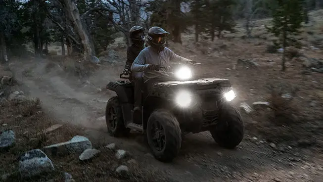 Two riders on a Polaris Sportsman Touring 850 ATV with illuminated headlights on a trail.