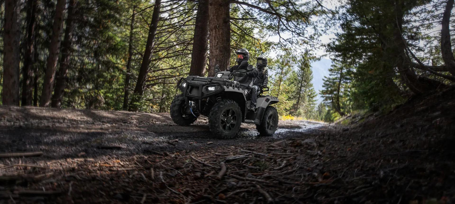 2 riders on a Polaris Sportsman Touring XP 1000 Trail ATV driving on a forest trail.