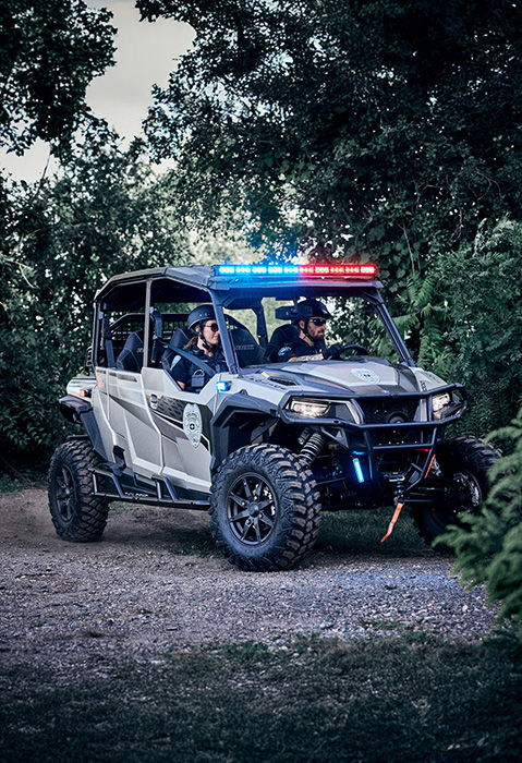 2 police officers riding in the 6-seat fully enclosed cab of police UTV with illuminated red and blue lights while driving driving on a dirt path through dense green foliage.