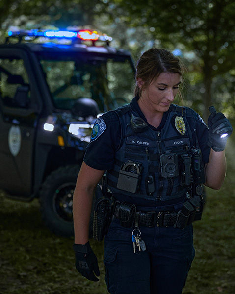 A female police officer, stands in a dimly lit, wooded area, holding a lit flashlight in her gloved hand. Behind her, a Polaris RANGER UTV with flashing red and blue emergency lights illuminates the background.