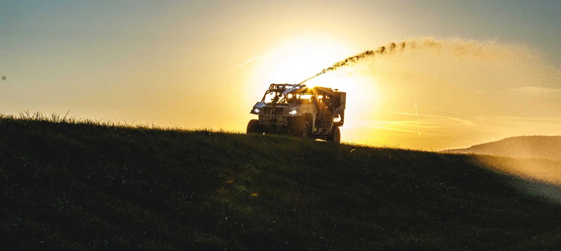 Polaris Government and Defense DAGOR military vehicle spraying water against a vibrant sunset over a grassy hill.