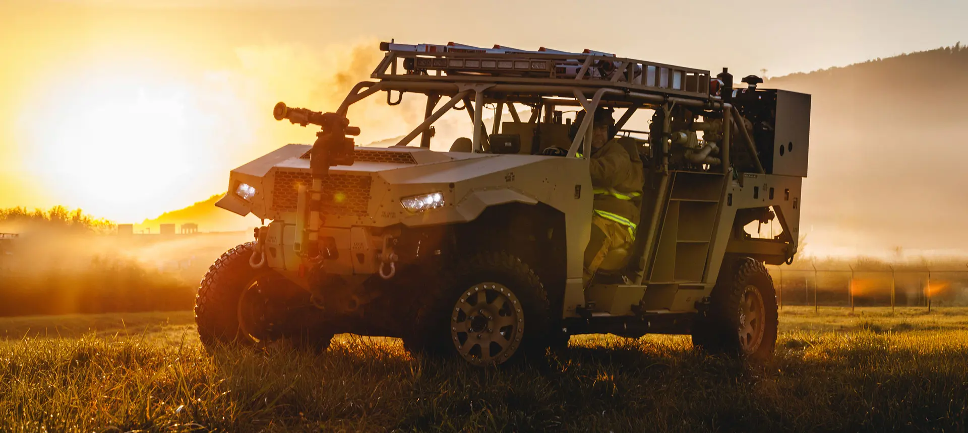 Polaris Government and Defense DAGOR light tactical vehicle with a ladder on its roof, parked in a misty field during sunrise, with a driver visible.