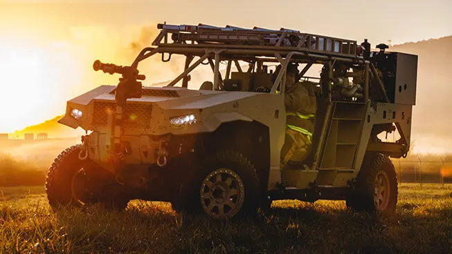 Polaris Government and Defense DAGOR light tactical vehicle with a ladder on its roof, parked in a misty field during sunrise, with a driver visible.