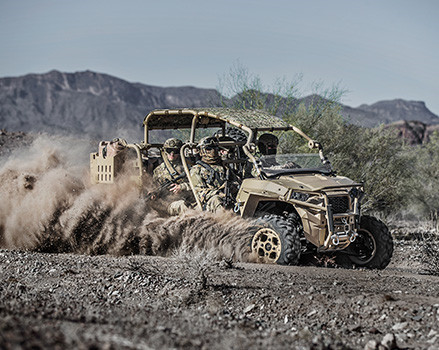 A tan MRZR Diesel light tactical vehicle with four military personnel kicks up a large cloud of dust while driving through a arid, rocky landscape.