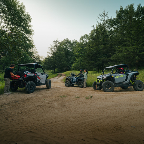Three Polaris Off Road vehicles on a dirt trail in a forest, with two people standing beside the lead vehicle.