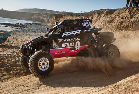 A Polaris RZR vehicle with the #1854 racing in a dusty terrain, showcasing sponsors like Yokohama and Optima.