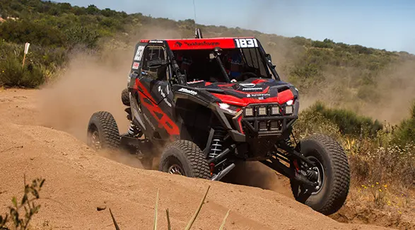 Côte à côte Polaris RZR Pro XP 2025 prenant un virage sur un sentier poussiéreux et projetant du sable.