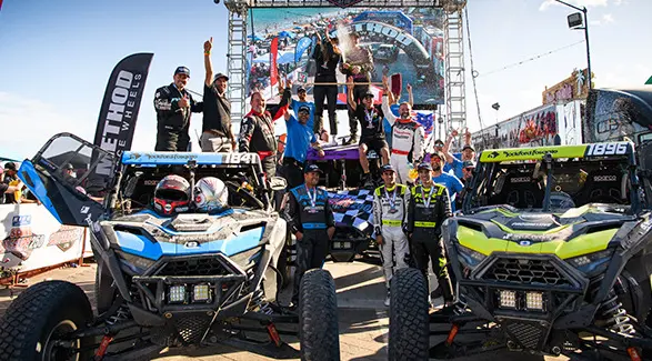 The RZR Racing team celebrating with trophies around two RZR vehicles at the finish line of a race under clear skies.