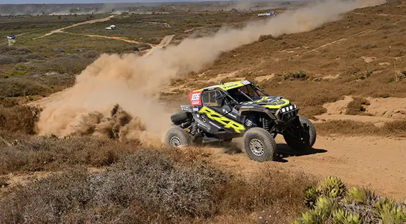 2025 Polaris RZR Pro XP racing on a dirt track, kicking up a large cloud of dust in a dry, grassy area with hills in the background.