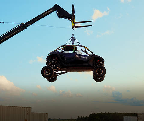 A black and blue RZR vehicle is suspended in mid-air by cables attached to a crane. The vehicle is set against a clear sky with a few clouds, and the crane arm extends diagonally from the top left corner of the frame.