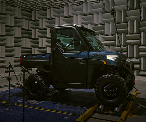 A Polaris RANGER UTV is positioned on a platform in a sound testing facility. Microphones on stands are placed around the vehicle to measure its noise levels or sound profile.