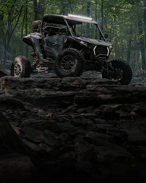 A black and red Polaris RZR side-by-side vehicle is parked in front of a large stack of cut logs and green trees.