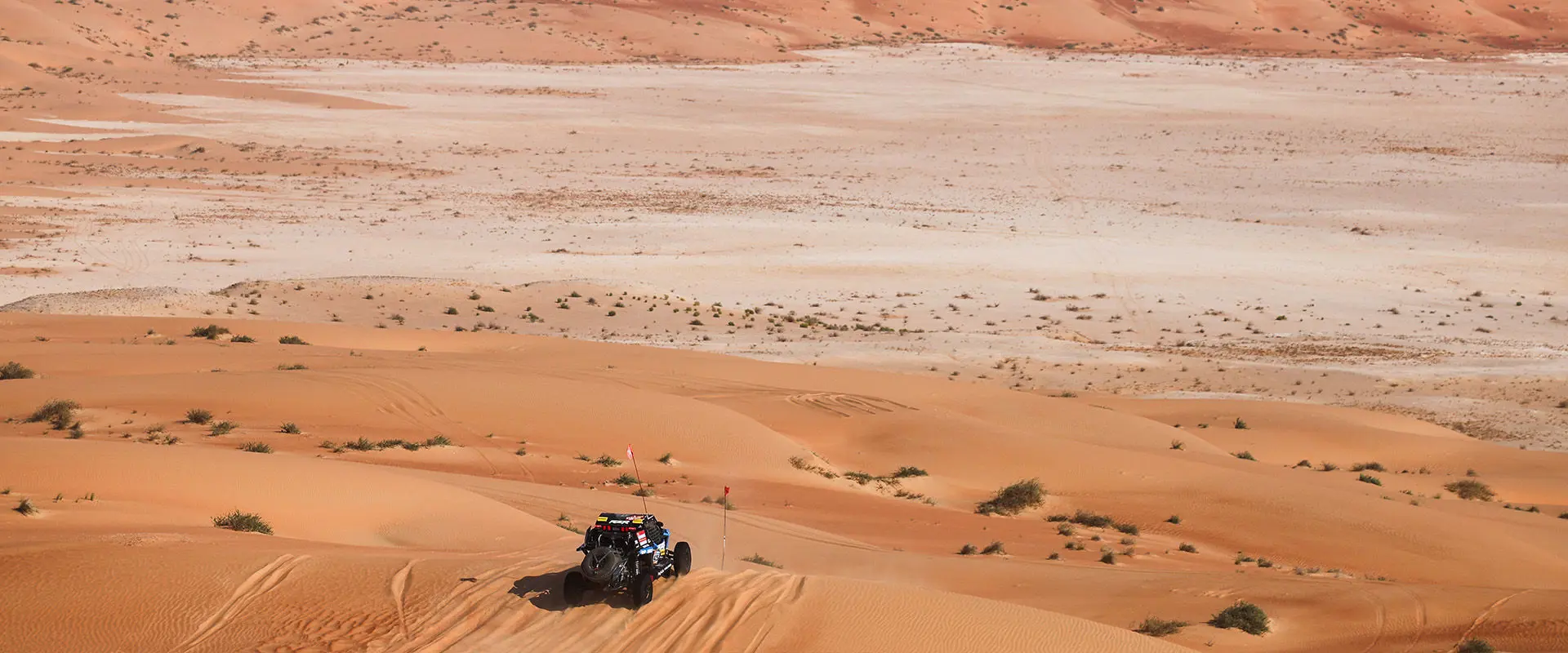Dakar Rally RZR Pro R Factory driving across a sandy dune landscape with two flags.