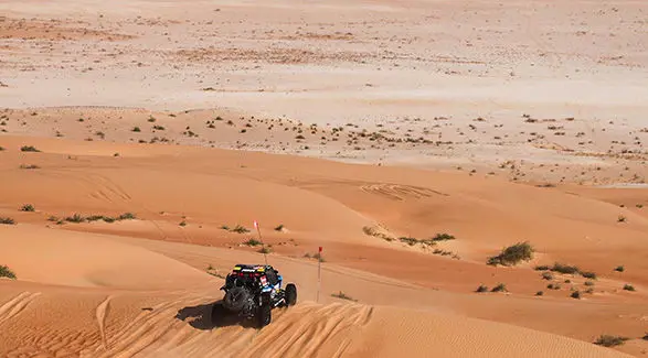 Dakar Rally RZR Pro R Factory driving across a sandy dune landscape with two flags.