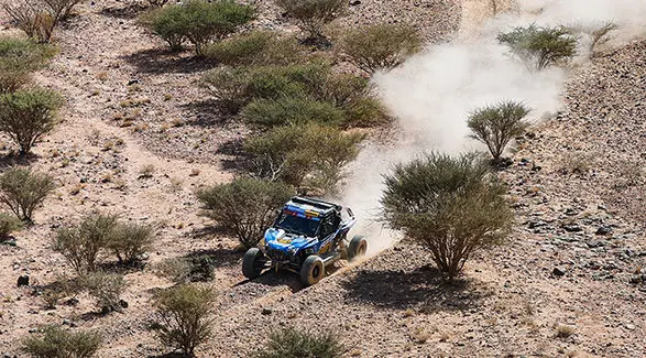 Dakar Rally RZR Pro R Factory driving through rough desert terrain with trees.