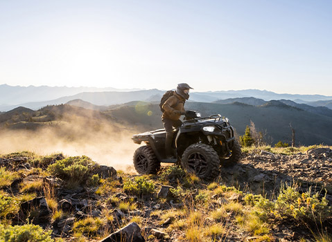 A person in a helmet and gear rides a Polaris Sportsman ATV up a dusty, rocky hill with mountains in the background.