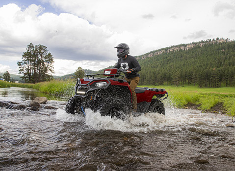 A person in a helmet rides a red Polaris Sportsman ATV through a shallow, rocky stream in a lush, green forest.
