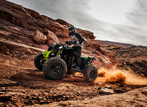 A person in a helmet rides a green Polaris Sportsman ATV up a dusty, rocky trail in a desert landscape.